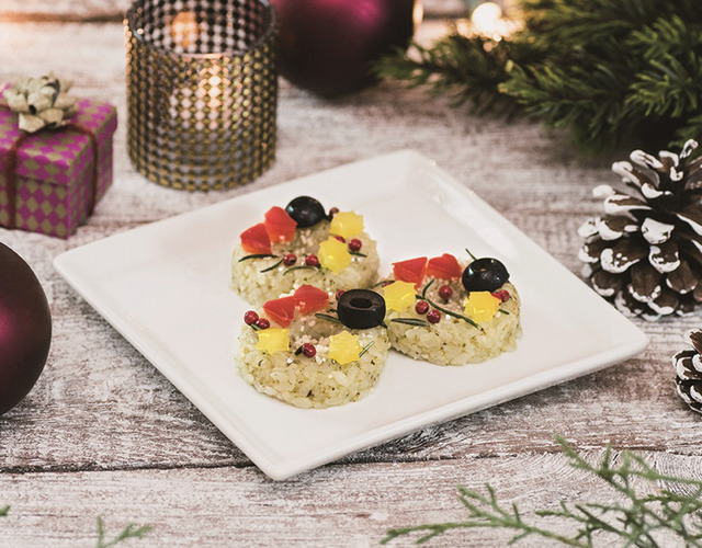 オニギリスタンド ギュッ(onigiri stand Gyu!)によるクリスマスリースをイメージした輪っか状のバジル風味おにぎりに2種類のチーズとオリーブ、パプリカ、ピンクペッパーなどをトッピングした「クリスマスリース風 チーズ&バジルおにぎり」(税込378円)